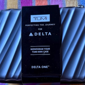 May include: A dark gray Tumi mini case with a textured, ridged design. A black label displays the Tumi logo, the Delta logo, and the text "Perfecting the Journey for Delta." The label also reads "Monogram Your Tumi Mini Case" and "Delta One."