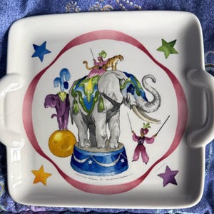 Puede incluir: Bandeja de cerámica blanca con un diseño de elefante de circo. La bandeja presenta un gran elefante gris con un tigre y artistas. El diseño incluye una cinta rosa, estrellas y un elefante morado sobre una bola amarilla.