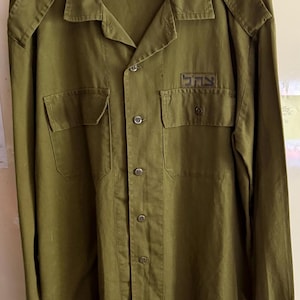 Puede incluir: Camisa verde oliva de estilo militar con dos bolsillos en el pecho y charreteras. La camisa tiene mangas largas y cuello. Un pequeño parche con texto hebreo es visible en el bolsillo derecho del pecho.
