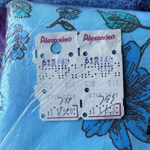 K&ouml;nnte beinhalten: Ein Paket blauen Stoffes mit einem Blumenmuster, das gro&szlig;e blaue Blumen und gr&uuml;ne Bl&auml;tter zeigt. Zwei Preisschilder sind sichtbar, mit dem Text "Alexander's" und verschiedenen Zahlen und Codes.
