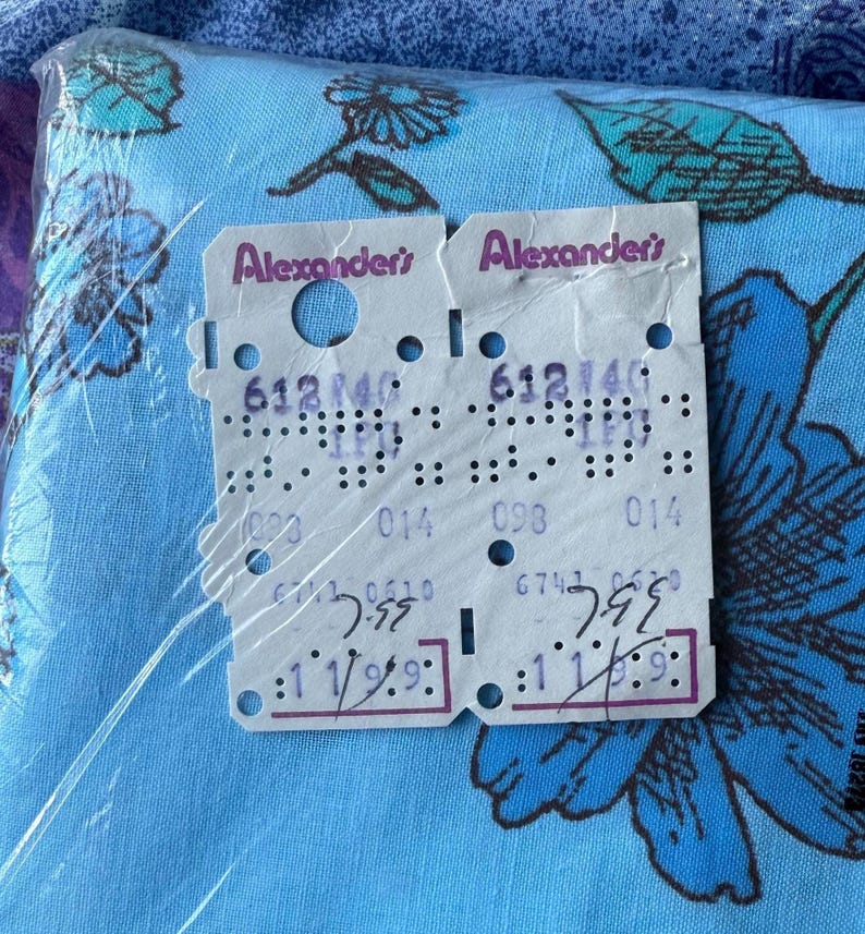 K&ouml;nnte beinhalten: Ein Paket blauen Stoffes mit einem Blumenmuster, das gro&szlig;e blaue Blumen und gr&uuml;ne Bl&auml;tter zeigt. Zwei Preisschilder sind sichtbar, mit dem Text "Alexander's" und verschiedenen Zahlen und Codes.