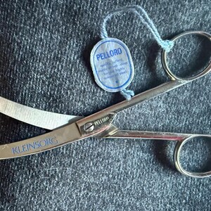 Könnte beinhalten: Edelstahlschere mit dem blauen Text "KLEINSORG". Die Schere hat ein Etikett mit der Aufschrift "PELLORO". Die Schere liegt auf einer dunkelgrauen, strukturierten Oberfläche.