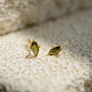 May include: A pair of gold stud earrings featuring marquise-cut olive green gemstones. Each earring is secured with a delicate gold prong setting, showcasing the elongated, pointed shape of the stones. The text GEMICA is visible in the corner.