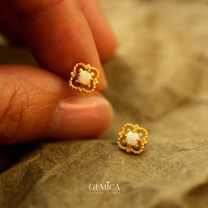 Puede incluir: Primer plano de dos pendientes de botón dorados. Cada pendiente presenta un diseño floral de cuatro pétalos con una pequeña piedra de ópalo blanca y redonda en el centro. Los pendientes están sostenidos por los dedos de una persona. La marca "GEMICA Jewelry Studio" está en la parte inferior.