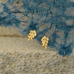 May include: A pair of gold-colored leaf-shaped stud earrings. The earrings are displayed on a textured white and blue surface. The text "SUMMER GARDEN JEWELRY STUDIO" is visible in the background.