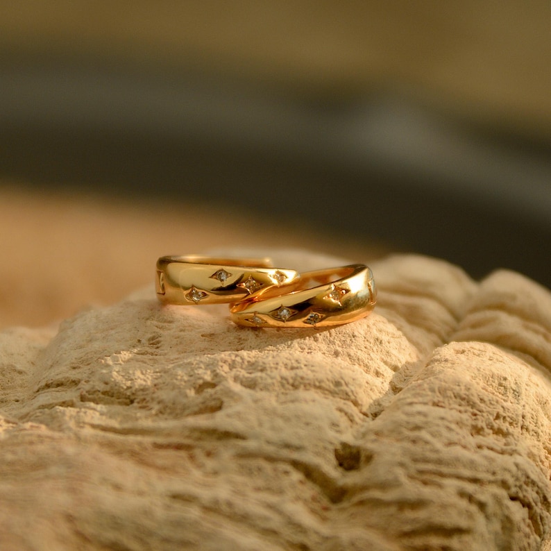May include: Two gold rings with small diamond accents. The rings are set on a light brown, textured surface. The rings have a polished finish and are reflecting the light.