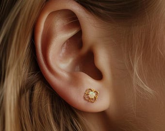 Puede incluir: Primer plano de un pendiente de oro con un diseño cuadrado de filigrana. El pendiente presenta una pequeña gema redonda de color blanquecino en el centro. Se ve una oreja, con pelo rubio. Joyería.