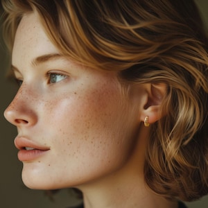May include: A side profile of a person with fair skin, freckles, and short, wavy blonde hair. The person is wearing small, gold hoop earrings. The background is a muted, neutral color.