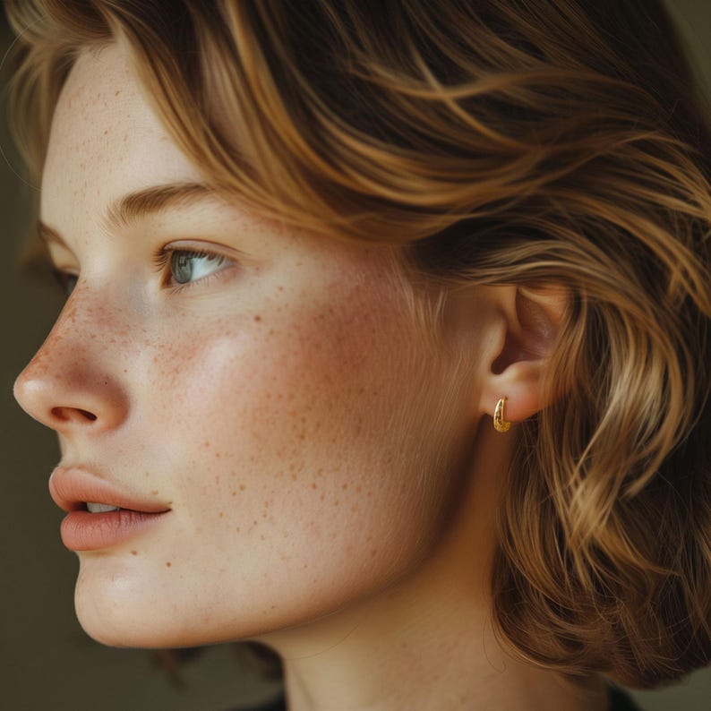May include: A side profile of a person with fair skin, freckles, and short, wavy blonde hair. The person is wearing small, gold hoop earrings. The background is a muted, neutral color.