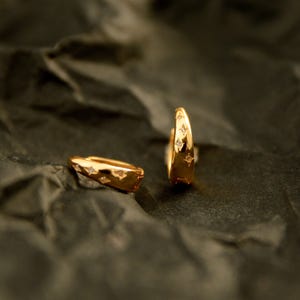 May include: Two gold-colored hoop earrings with star-shaped cutouts and small, clear gemstones. The earrings are displayed on a dark, textured surface, showcasing their polished finish and intricate design. The earrings are a jewelry item.