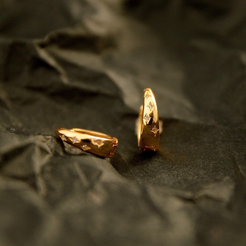 May include: Two gold-colored hoop earrings with star-shaped cutouts and small, clear gemstones. The earrings are displayed on a dark, textured surface, showcasing their polished finish and intricate design. The earrings are a jewelry item.