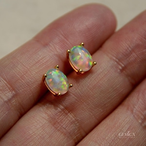 May include: A pair of oval-shaped opal stud earrings set in gold metal prongs. Each opal displays a milky white base with iridescent flashes of green, orange, and pink. The text GEMICA is printed in the bottom right corner of the image.