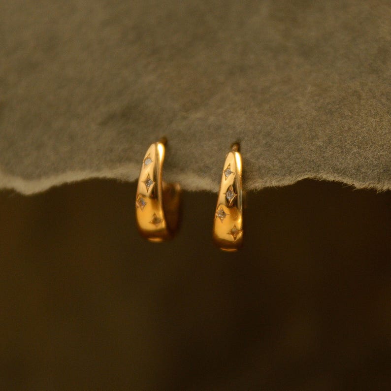 May include: A pair of gold-colored hoop earrings. Each earring features a series of small, sparkling diamond-like accents. The earrings have a smooth, polished finish and a curved design, creating a simple yet elegant look.