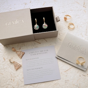 May include: An open jewelry box with a pair of hoop earrings, rings, and a jewelry pouch. The earrings have a clear orb with colorful inclusions. The rings are gold-toned. A card with care instructions is also visible.