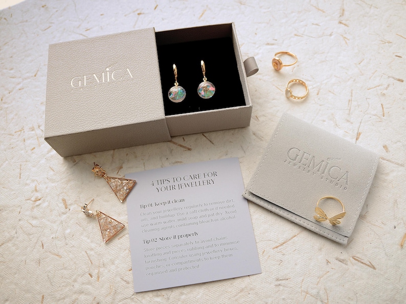 May include: An open jewelry box with a pair of hoop earrings, rings, and a jewelry pouch. The earrings have a clear orb with colorful inclusions. The rings are gold-toned. A card with care instructions is also visible.