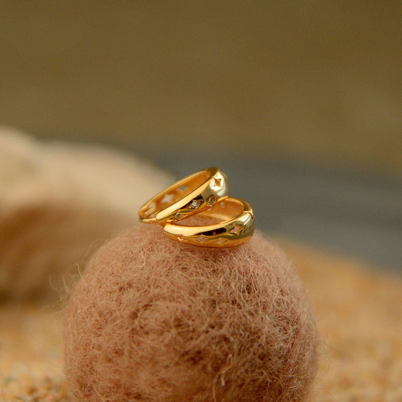 May include: A pair of gold-colored hoop earrings with star-shaped cutouts and small, clear gemstones. The earrings are displayed on a textured, brown surface, with a soft, blurred background.