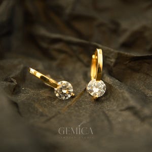 Peut inclure: Une paire de boucles d'oreilles créoles dorées, chacune ornée d'une seule pierre précieuse ronde et transparente. Les boucles d'oreilles sont présentées sur un fond sombre et texturé. La marque "GEMICA JEWELRY STUDIO" est imprimée en bas.