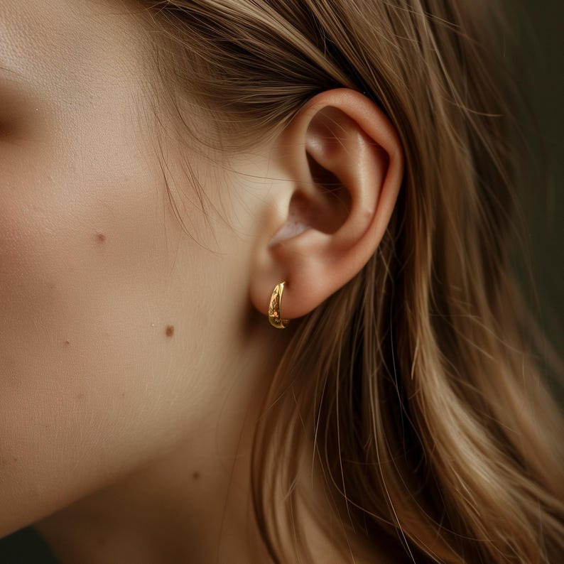 May include: Close-up of a gold hoop earring on an ear. The earring has a textured surface and a small, curved design. The person's hair is long and blonde, with a soft, blurred background.