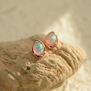 May include: A pair of opal stud earrings. Each earring has a pear-shaped, iridescent opal stone set in a rose gold bezel. The opals show a mix of pink, blue, and green hues. The earrings are displayed on a light brown, textured surface.