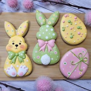 May include: An assortment of Easter-themed cookies on a wooden board. The cookies include two bunny shapes, one yellow and one green, and two egg shapes, one yellow and one pink. The cookies are decorated with colorful icing and polka dots.
