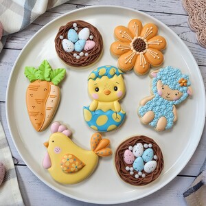 May include: A white plate displays Easter-themed biscuits. The biscuits include a carrot, a chick hatching from an egg, a lamb, a flower, two nests with eggs, and a chicken. The biscuits are decorated with orange, yellow, blue, and green icing.
