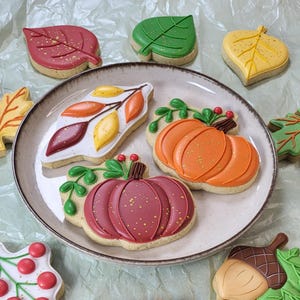 May include: A plate of decorated fall-themed cookies. The cookies are shaped like leaves, pumpkins, and an acorn. The leaves are red, green, and yellow. The pumpkins are orange and burgundy. The cookies are decorated with icing and gold sprinkles.