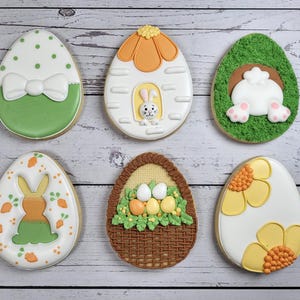 May include: Six Easter egg-shaped biscuits with detailed icing designs. The biscuits feature various themes, including a bow, a bunny in a house, a bunny's rear, a bunny with carrots, a basket of eggs, and a floral design. The biscuits are on a light wood surface.