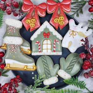 May include: A collection of decorated Christmas cookies on a dark plate. The cookies include ice skates, mittens, a gingerbread house, a reindeer, and red bows with gold accents. The cookies are decorated with green, white, red, and gold icing.
