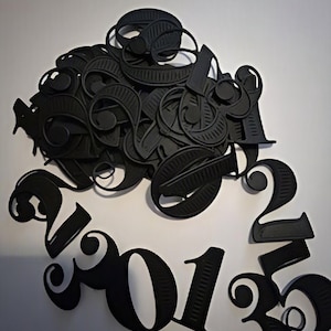 May include: A collection of matte black, three-dimensional plastic numbers featuring a decorative, serif-style font. The pile includes various digits such as 0, 1, 2, 3, and 5, with some arranged in a curved formation at the bottom of the cluster.