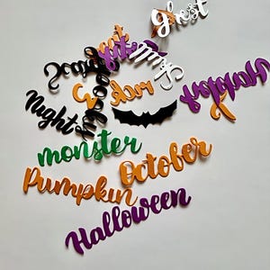 Può includere: Ritagli a tema Halloween assortiti in arancione, viola, nero, bianco e verde. I ritagli includono parole come "Halloween", "Ottobre", "Zucca", "Mostro" e "Fantasma". È presente anche un ritaglio di pipistrello nero.