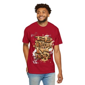 May include: A red t-shirt with the words "BE JOYFUL IN HOPE" in a graffiti-style design. The text is in gold and white, with a splatter effect. The shirt is short-sleeved.