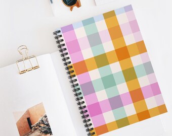 May include: A spiral-bound notebook with a colorful plaid pattern in shades of pink, orange, yellow, green, and purple. The notebook is open on a white surface, with a gold paper clip and sunglasses nearby.