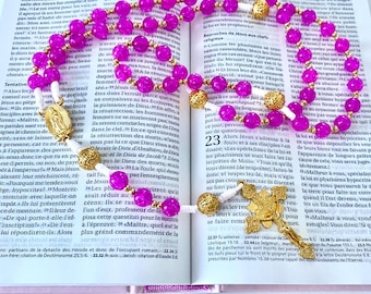 May include: A rosary with bright pink beads and gold-colored accents, including a crucifix and a centerpiece with a religious figure, rests on an open book. The book's pages display printed text.