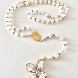 Peut inclure: Un chapelet blanc avec un pendentif en forme de croix dorée. Le chapelet est orné de perles blanches, de perles et d'accents dorés. La croix est ornée de cristaux transparents et de perles, et un médaillon doré est également présent.