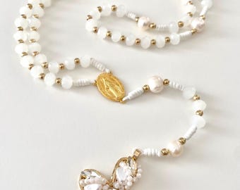 May include: A white rosary with a gold cross pendant. The rosary features white beads, pearls, and gold accents. The cross is adorned with clear crystals and pearls, and a gold medallion is also present.
