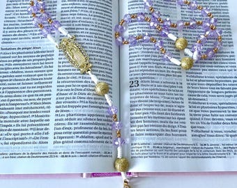 May include: A rosary with purple beads, gold accents, and a gold cross, resting on an open book. The beads are strung together, and the cross is detailed with clear stones. The book's pages are visible.