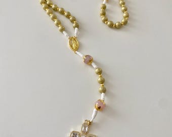 May include: Gold-colored rosary with pink beads and a crystal-studded cross pendant. The rosary features gold-colored, textured beads and white string. The cross is adorned with clear, rectangular crystals. A gold-colored medallion is also visible.