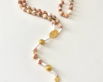 May include: A rosary with pink and brown stone beads, gold-colored accents, and a gold crucifix. The beads are strung on white cord, with gold-colored metal beads separating the decades. The crucifix features a detailed depiction of Jesus.