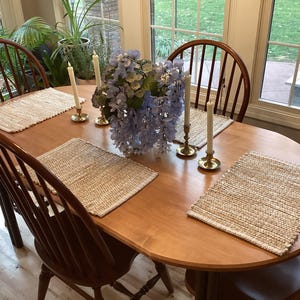 May include: A wooden oval dining table set for four with woven placemats, wooden chairs, candles in brass holders, and a blue floral centerpiece. The table is in a well-lit dining room.