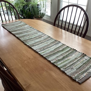 May include: A woven table runner with a striped pattern in shades of green, brown, and beige. The runner is rectangular and placed on a light-colored wooden table. Two wooden chairs are visible on either side of the table.