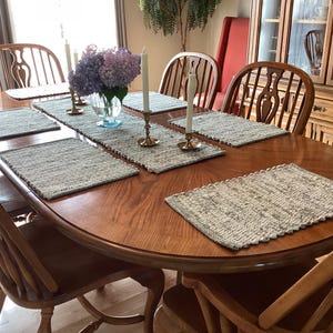 May include: A wooden dining table set with six chairs, featuring a long runner and matching rectangular placemats in a textured grey weave. A clear glass vase holds purple hydrangeas, accompanied by two brass candlesticks with white taper candles.