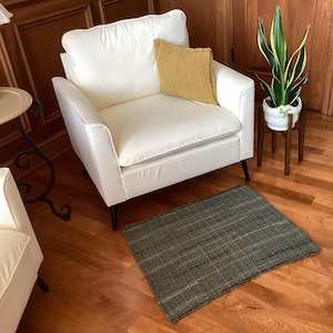 May include: A white armchair with a yellow throw pillow sits on a dark gray woven rug in front of a wooden wall. A potted plant with green leaves is on a wooden stand to the right of the chair.