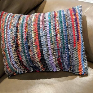May include: A colorful woven throw pillow with a striped pattern in shades of blue, red, orange, purple, and green. The pillow is made of recycled fabric and has a rustic, handmade look.