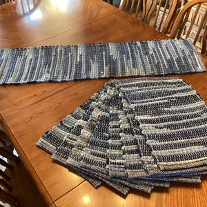 May include: A set of woven placemats and a matching table runner. The placemats are stacked in a fan shape, and the runner is rectangular. Both are made of blue, white, and gray woven fabric. The items are displayed on a wooden table.