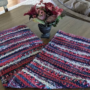 May include: Two handmade, rectangular rag rugs with a striped pattern in red, white, and navy blue. The rugs are made of woven fabric strips and are displayed on a wooden table. A decorative vase with flowers sits between them.