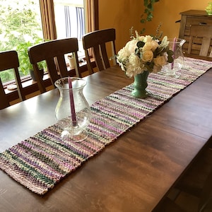 May include: A handwoven table runner in shades of purple, green, and tan, displayed on a dark wood dining table. The runner is decorated with a vase of white flowers and a glass candle holder with a purple candle. The table is set for a meal.