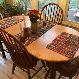 May include: A wooden dining table set with four chairs, two placemats, and a table runner. The table is set with candles and a vase of flowers. The placemats and runner have a red, green, and white woven pattern.