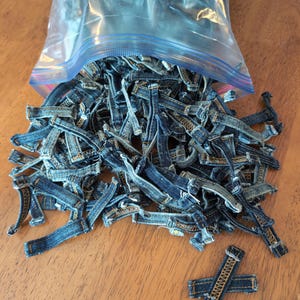 May include: A clear plastic bag filled with small, rectangular denim pieces, spilling onto a wooden surface. The denim pieces are various shades of blue with visible stitching, suggesting they are repurposed from jeans.
