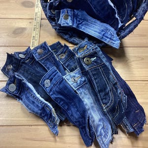May include: A collection of denim jacket pieces, including buttoned plackets and sections of the garment, in various shades of blue. The pieces are arranged on a wooden surface, suggesting a craft or upcycling project.