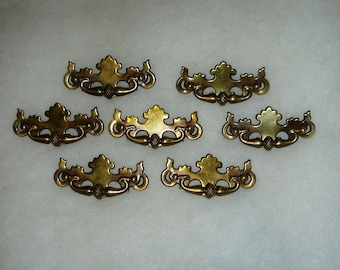 May include: A set of six brass-toned metal drawer pulls, each featuring a decorative, ornate backplate with a central acorn-shaped detail and curved, symmetrical scrollwork arms extending outward to circular ends on both sides.